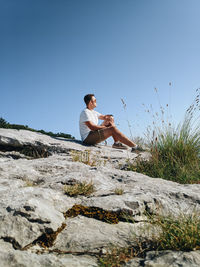 Young man sitting on rock against clear sky