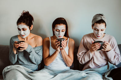 Female friends with facial masks holding mugs while sitting against wall at home