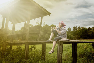 Full length of man playing flute while sitting on boardwalk over grassy field