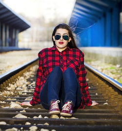 Portrait of beautiful woman wearing sunglasses while sitting on railroad track