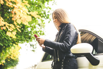 Trendy blonde girl using smartphone leaning against her car in a beautiful autumn afternoon. 