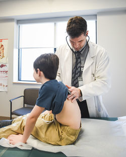 Doctor with stethoscope on a child's back on a clinic exam table.