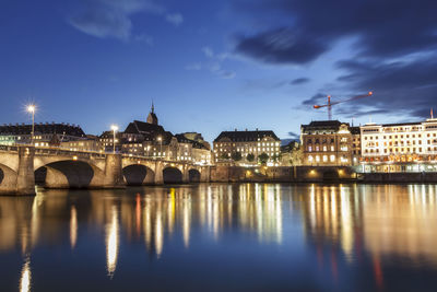 Illuminated bridge over river by buildings against sky at night