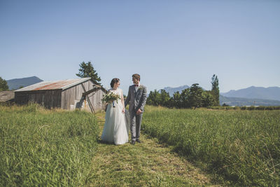 Newlywed couple looking face to face while walking on grassy field against barn and clear sky