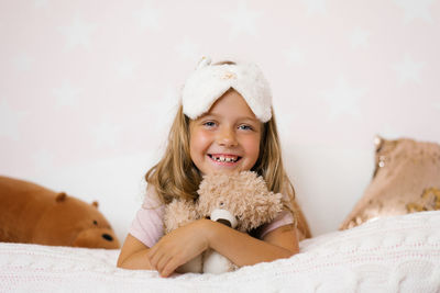 Portrait of a happy baby girl with a sleep mask on her head, lying on the bed and hugging 