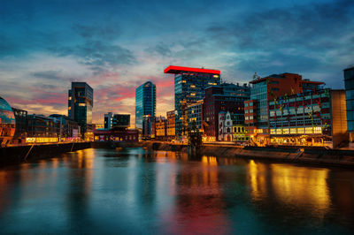 Illuminated buildings by river against sky at dusk