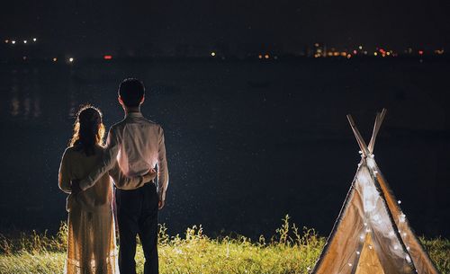 Rear view of couple standing by illuminated tent against river at night