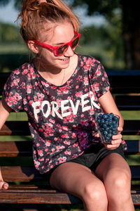 Smiling teenage girl wearing sunglasses while sitting blueberries on bench