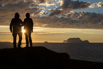 Rear view of silhouette friends standing on rock by sea against sky during sunset