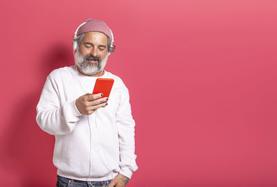 Man with hat and white beard listening to streaming music with headphones isolated on pink studio 