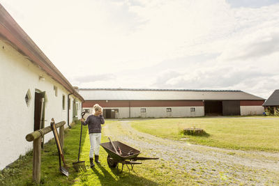 Woman using phone while holding rake against barn at farm