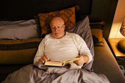 Senior man reading book while lying on bed at home