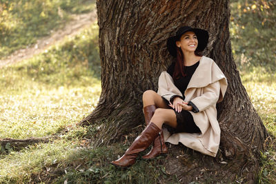 Smiling woman wearing hat sitting by tree at park