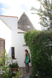 Woman standing by house against trees and plants