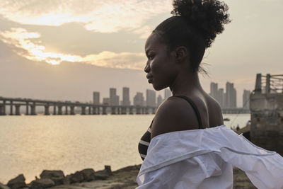 Tranquil pensive young african american female in casual clothes relaxing on urban seafront at sunset time