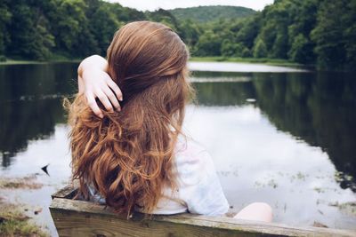 Rear view of woman with hand in hair sitting on bench at lakeshore