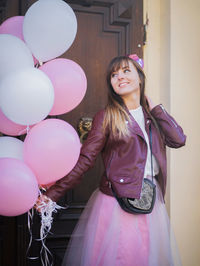 Smiling young woman holding pink balloons while standing against door