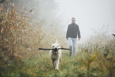 Mid adult man with dog walking on grassy field during foggy weather