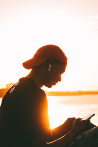 Side view of young man sitting at ocean using mobile phone against clear sky