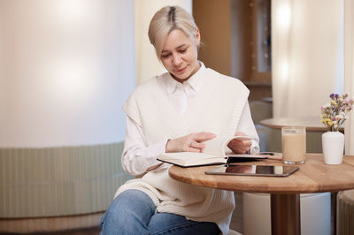 A cute girl in a shirt is flipping through a diary lying next to a tablet and a cup of coffee 