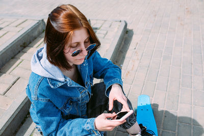Young stylish woman walks on street with a skateboard and uses a smartphone