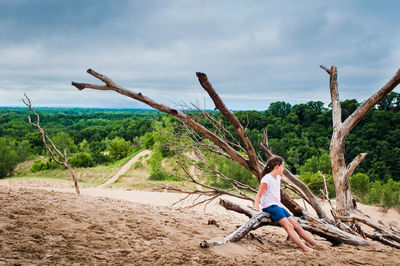 Side view of girl sitting on bare tree against forest
