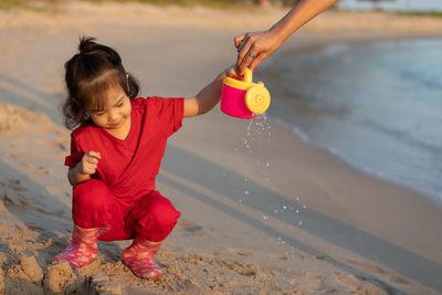 Cropped hand of mother giving watering can to daughter crouching at beach