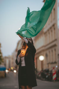 Portrait of beautiful woman moving green scarf while holding illuminated jar in city at dusk