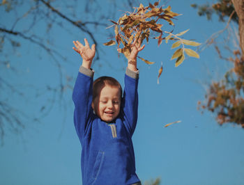 Smiling boy throwing dry leaves while standing against blue sky