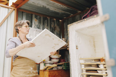 Potter with plank standing by rack at workshop