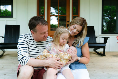A young girl holds her newborn baby sister with her parents on a porch
