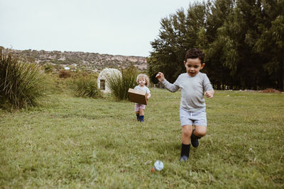 Boy running with male friend carrying cardboard box while playing on meadow