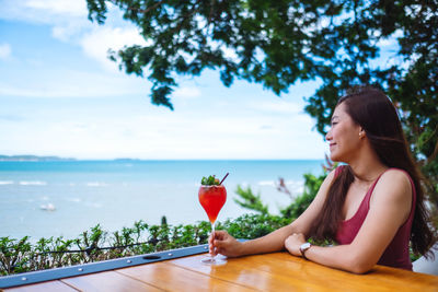 Portrait of young woman holding wineglass by sea against sky