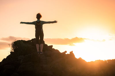 Rear view of woman with arms outstretched standing on cliff against orange sky