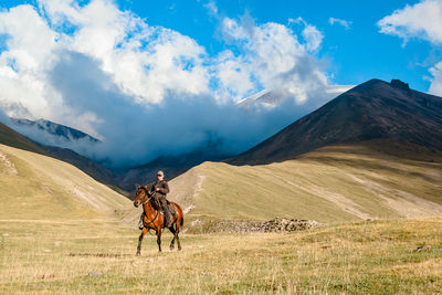 A cowgirl shepherd rider on a horse gallops through a mountain valley on a clear day against sky