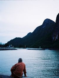 Rear view of man sitting on riverbank against clear sky