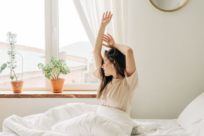 Young  girl stretches in the morning after waking up while sitting on the bed in a cozy bedroom.