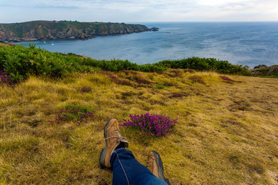 Low section of man sitting on grassy field by sea against sky