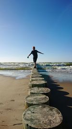 Boy balancing on tree stumps at beach against blue sky