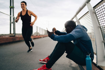 Sportsman holding mobile phone while cheering female athlete running on footbridge