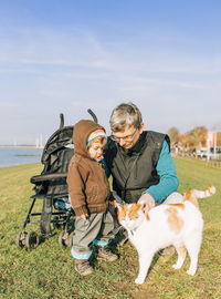 Grandfather with granddaughter looking at cat standing on grassy field against sky