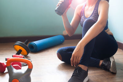 Low section of woman holding water bottle while sitting on wooden floor by sports equipment