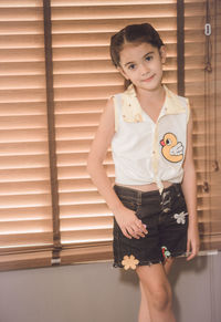 Portrait of cute smiling girl standing against window blinds