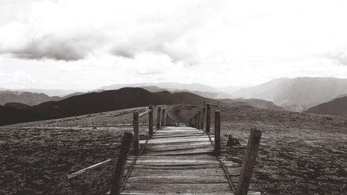 Empty wooden footbridge leading towards mountains against sky