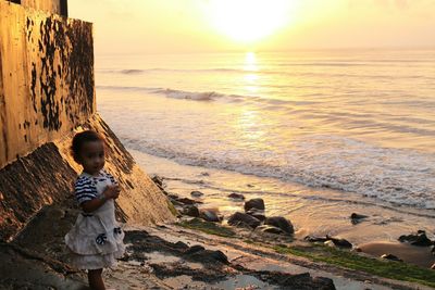 Boy standing on rock at beach against sky during sunset