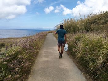 Rear view of man walking on footpath by sea against sky