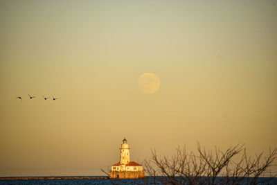 Silhouette birds flying against sky with moonrise over lighthouse during sunset