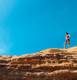 Low angle view of young woman in bikini standing on rock against clear blue sky
