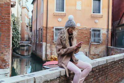 Young woman using mobile phone while sitting on retaining wall against canal and buildings in city