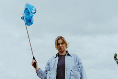 Low angle portrait of confident young woman collecting garbage against sky